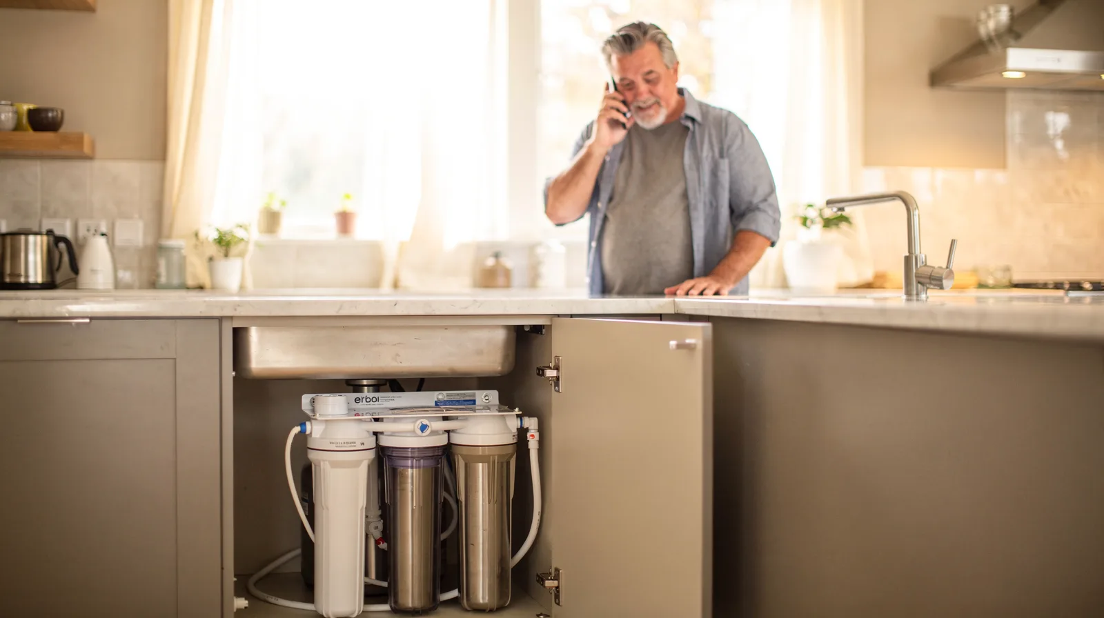 A homeowner answering a phone call in a kitchen beside a water filter system under the sink