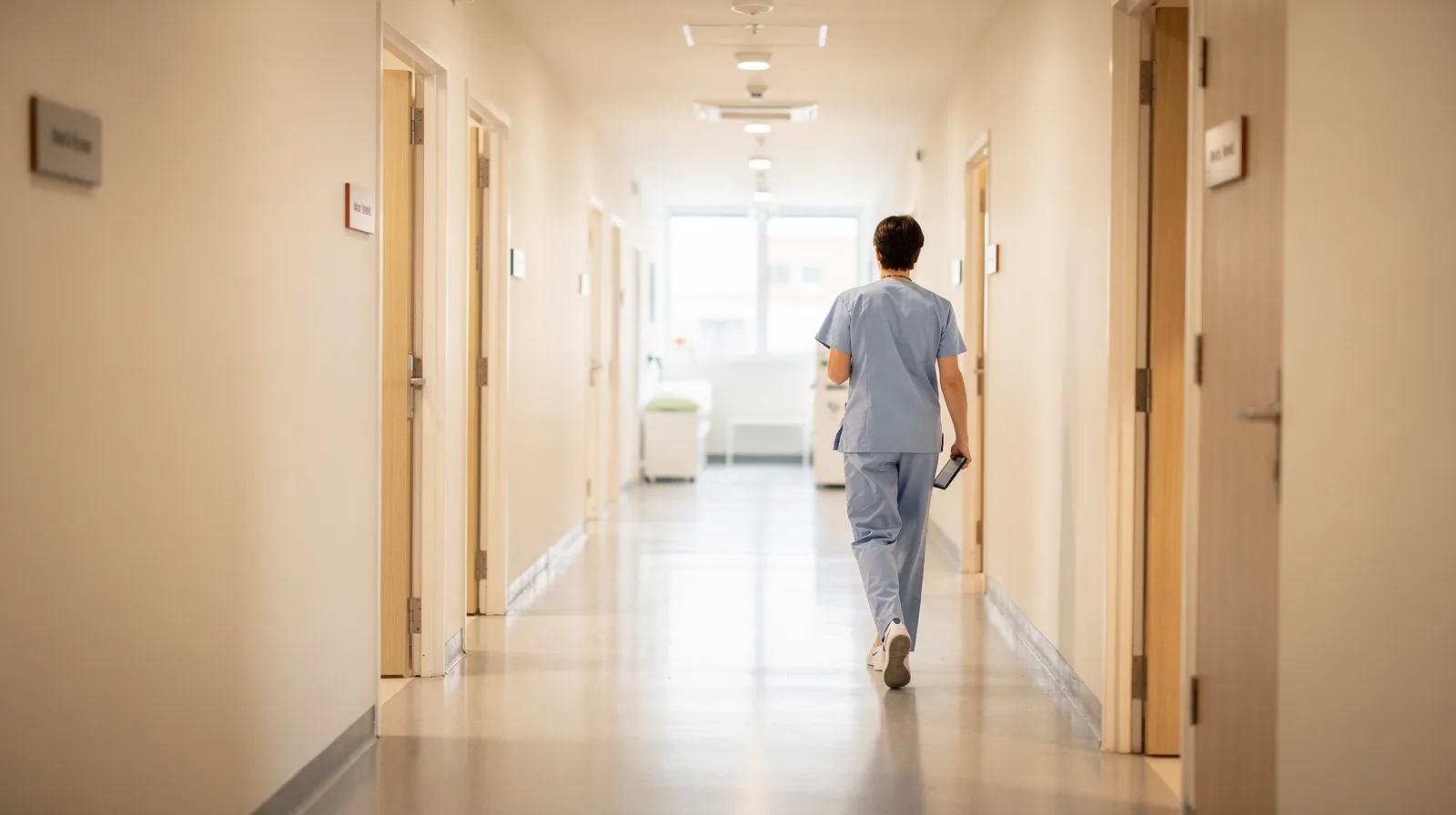 A clinic hallway with several exam room doors and a staff member walking with a tablet
